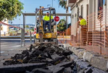 Máquina excavadora amarilla trabajando junto a una acera, con fragmentos de pavimento levantado en primer plano y una zona delimitada por una malla naranja.