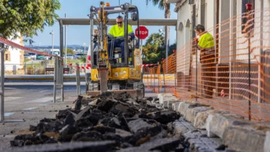 Máquina excavadora amarilla trabajando junto a una acera, con fragmentos de pavimento levantado en primer plano y una zona delimitada por una malla naranja.