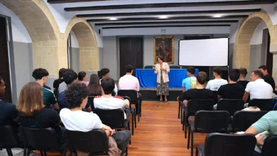 Una mujer habla ante un grupo de estudiantes sentados en filas de sillas en una sala con arcos de piedra y vigas oscuras en el techo.