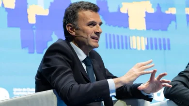 Hombre con traje oscuro y corbata azul sentado en un sillón blanco, gesticulando con ambas manos y utilizando un micrófono de diadema.