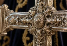 Primer plano de una cruz procesional de metal ornamentado junto a un tejido ceremonial, con el fondo desenfocado.