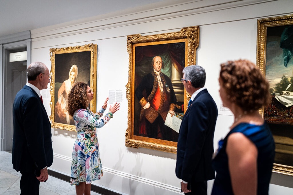 Dos personas conversan en una sala institucional con las banderas de Estados Unidos, España y la Unión Europea al fondo.