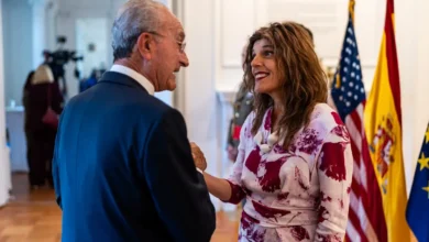 Un hombre con traje azul conversa con una mujer con vestido estampado en una sala donde aparecen las banderas de Estados Unidos, España y la Unión Europea.