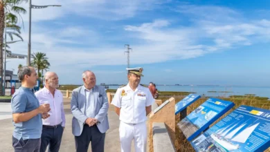 Cuatro hombres junto a varios paneles informativos azules sobre el buque escuela Juan Sebastián de Elcano, en un paseo exterior con el mar al fondo.