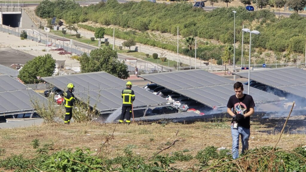 Pastos secos y piedras en primer plano, con humo blanco y gris elevándose detrás de una valla metálica.