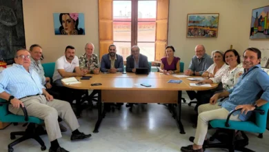 Grupo de personas sentado alrededor de una mesa de reuniones en una sala con ventanas de madera abiertas y cuadros en las paredes.