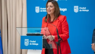 Mujer con chaqueta roja ante un atril transparente y un micrófono, con un fondo azul que muestra los logotipos de Málaga y Ciudad de Málaga.