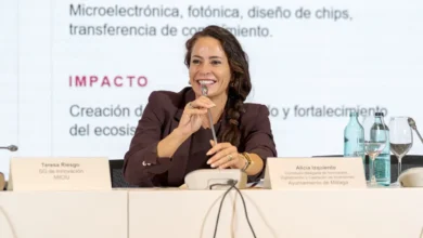 Una mujer con chaqueta granate interviene con un micrófono en una mesa de reunión; delante hay placas identificativas, botellas de agua y copas, y detrás aparece un panel con texto sobre tecnologías e impacto.
