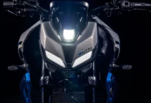 Vista frontal de una motocicleta Yamaha MT-125 con iluminación blanca encendida, carrocería gris, detalles azules y fondo negro.