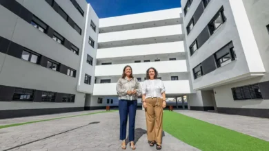 Dos mujeres frente a un conjunto residencial de cuatro plantas con fachadas blancas y grises, patio central y zonas ajardinadas.