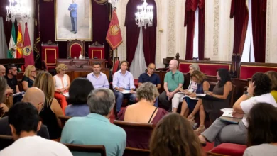 Reunión institucional con varias personas sentadas en un salón municipal con banderas, cortinas rojas y mobiliario oficial.