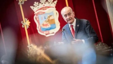 Hombre con traje interviene ante un micrófono en un salón institucional con cortinas rojas y un escudo al fondo